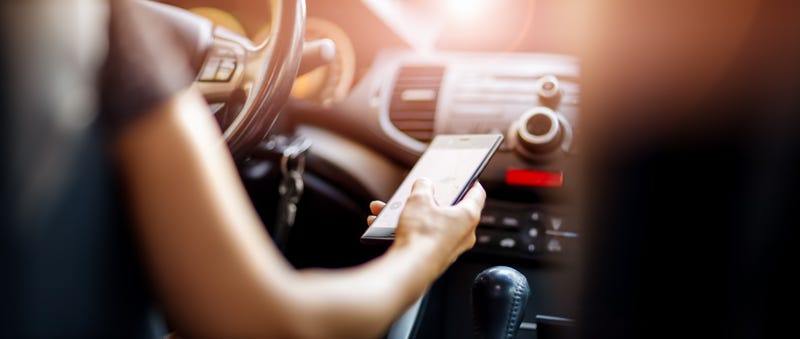 Driver driving a car on asphalt road in summer day at park. Woman at steering wheel with smartphone in her hand.