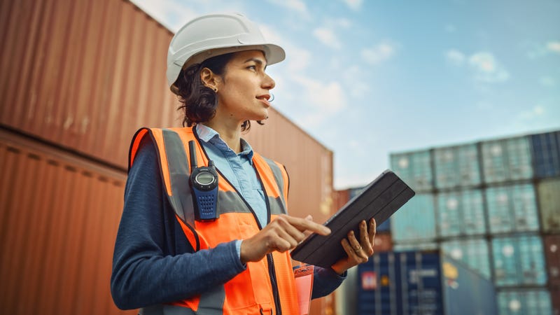 woman with hard hat, safety vest and tablet stands outside a building