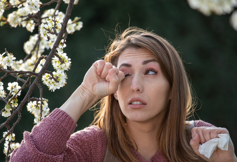 Young pretty girl rubbing eyes beside blooming tree in spring time. Itchy eyes from allergy