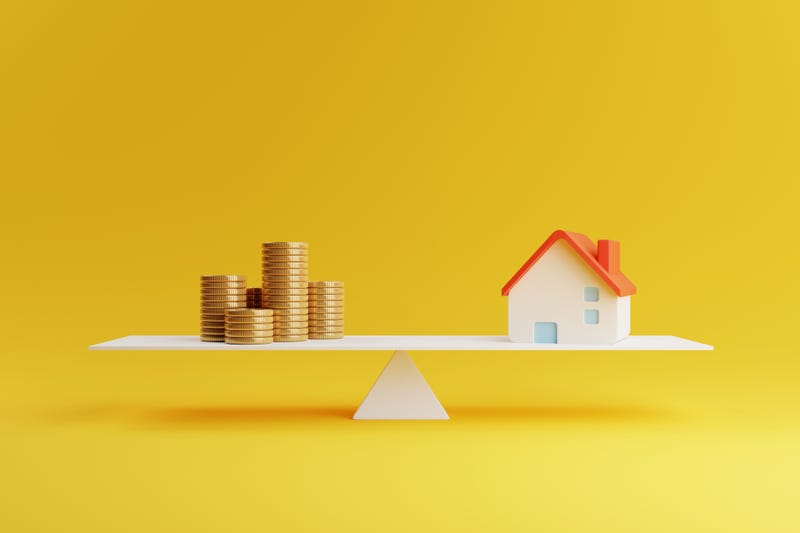 A little house and coins sitting on a balance beam. 