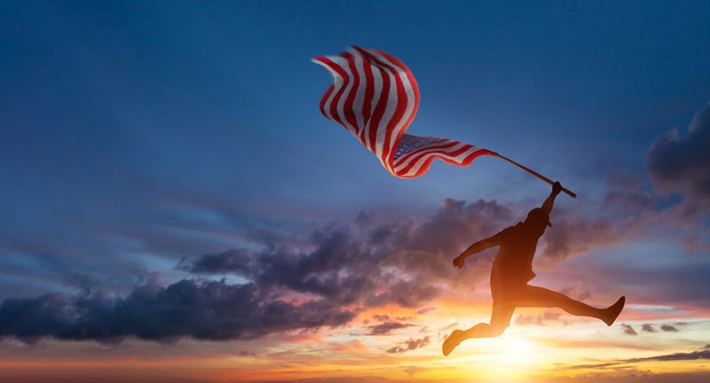 Silhouette of man jumping while holding US American flag in front of beautiful sunrise.