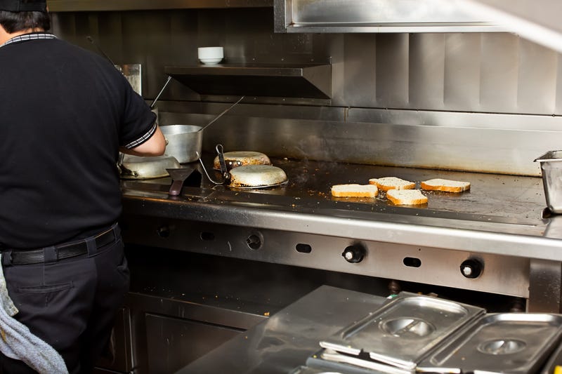 A view of a line cook attending a stove and griddle area. Slices of bread are toasting on the griddle, in a restaurant kitchen setting.