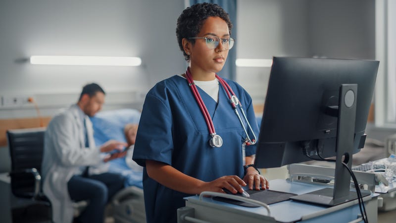 A nurse uses a computer while a doctor checks on a patient in the background.
