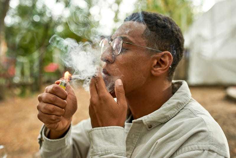 Close view of young afro guy lighting a joint, wearing grey shirt and glasses, smoking on the fresh air , a lot of smoke on the picture, holding a yellow lighter in hand