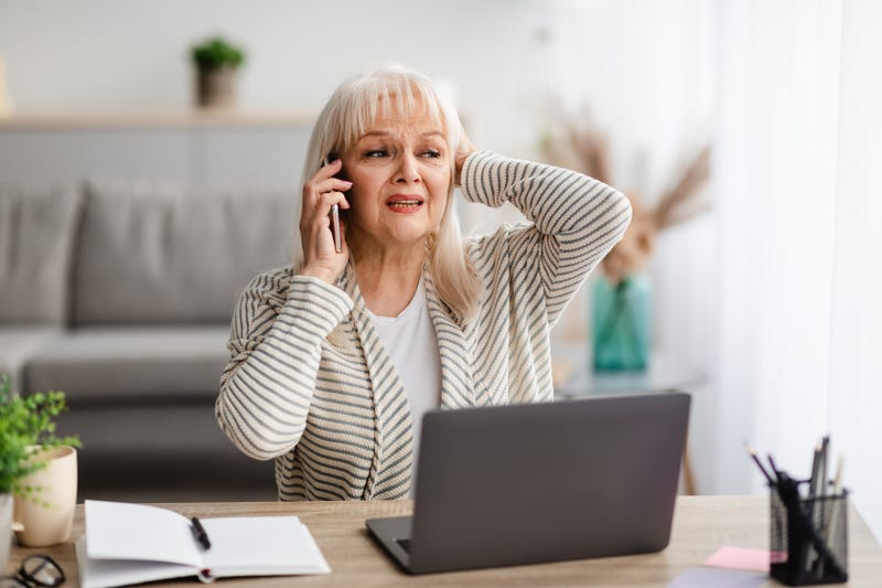 Portrait of stressed senior lady making call using mobile phone near window in modern office. Fustrated mature woman having unpleasant conversation, showing negative emotion, grabbing head