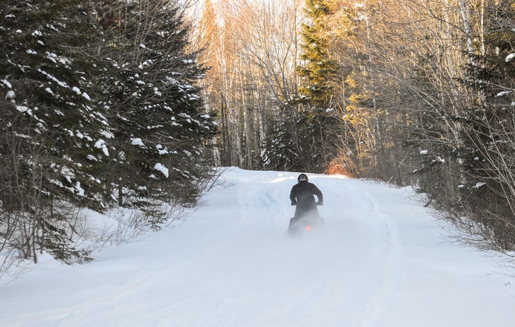 A snowmobiler seen driving away through a clearing in a wooded area.