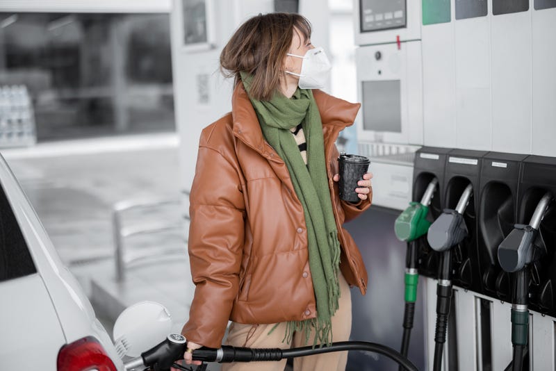 Woman refueling car with gasoline standing with a coffee and holding refueling gun at petrol station. Woman wearing face mask and warm clothes