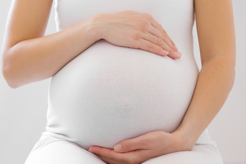 Young adult pregnant woman in white clothes touching big belly and feeling her baby move on light gray background. Pregnancy time concept. Expectation time. Closeup. Front view.