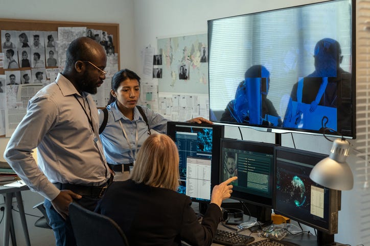 Three police officials huddle around four computer monitors