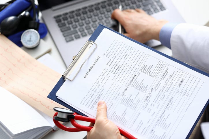 Close up of a doctor holding a clipboard with patient history documents, a stethoscope, and typing on a laptop.