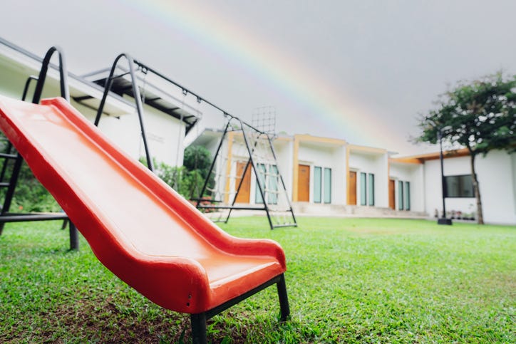 A slide and swing set sitting empty in front of a child care center with a rainbow in the back