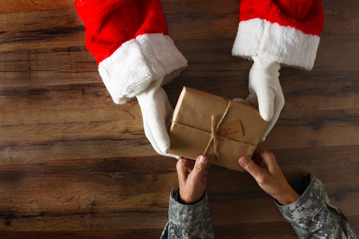 Photo of Santa handing a plainly wrapped present to a soldier. Only their hands holding the gift are visible.