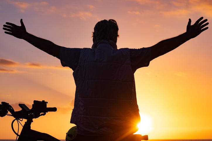 Mature man at the beach close to his bicycle enjoying sunset orange light with outstretched arms.