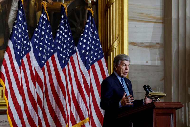 roy blunt speaking at a podium in front of four american flags