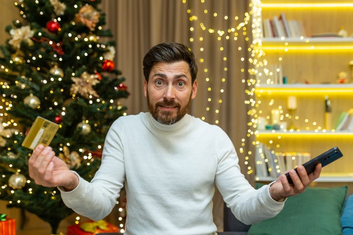 Dissatisfied and disappointed man looking at camera nervously trying to do online shopping while sitting for christmas and new year, holding bank credit card and smartphone.