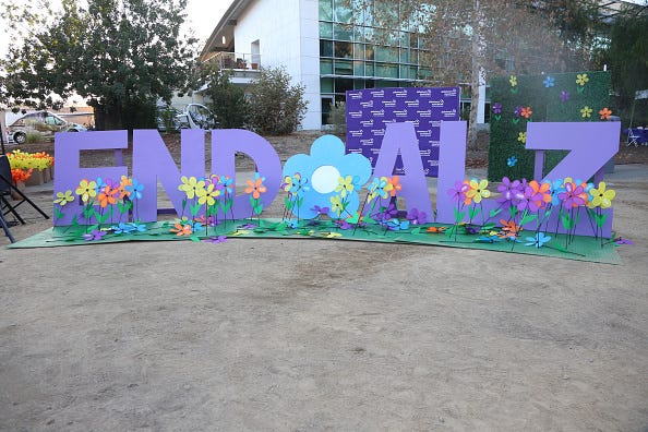 General atmosphere a Alzheimer's Association 2022 Walk to End Alzheimer's. Big purple block letters spell out "End ALZ" surrounded by yellow, orange, blue. and purple plastic flowers. An Alzheimer's Association step and repeat banner is in the background