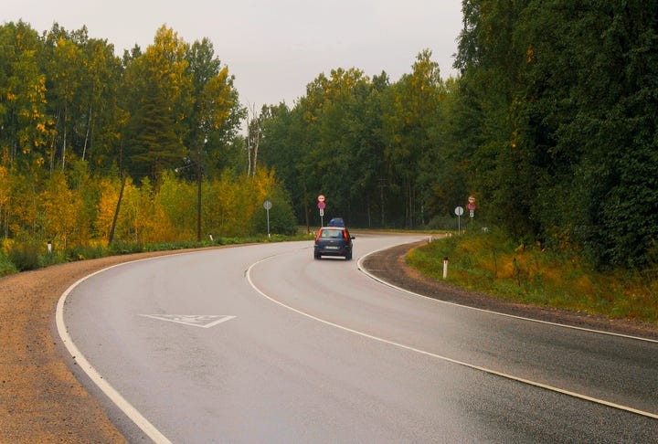 A two-lane rural highway with forested area on both sides. A couple of cars can be seen going around a curve.