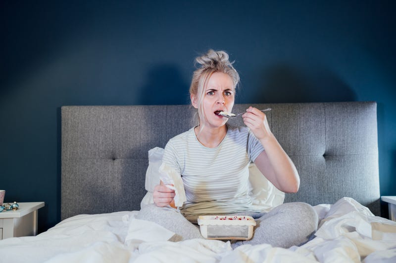 Shocked, stressed, surprised woman eating ice cream, holding a remote control while watching TV in bed. Scary horror movie or terrible news, scare in her eyes Overeating, Jamming of negative emotions.