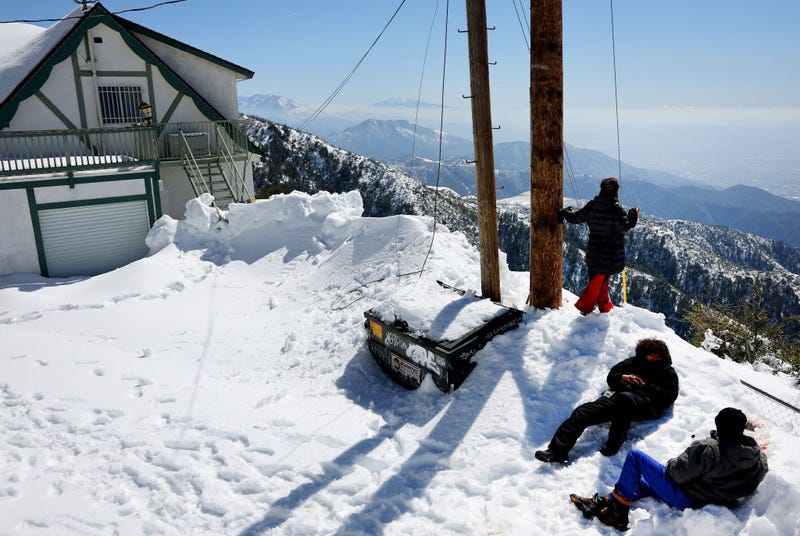 Dion Slider (R) waits in the snow with his wife Jennifer and son Kai, as they wait for their phones to charge at a closed restaurant (L), after a series of winter storms in the San Bernardino Mountains in Southern California on March 3, 2023 in Crestline, California. The Slider family lost power in their snowed-in home four days ago and walked to the restaurant to charge their phones, which has an outdoor electrical socket. Some residents have been stranded in Crestline more than a week due to the snowfall while the local grocery store was severely damaged when its roof collapsed under the weight of the snow. California Governor Gavin Newsom has declared a state of emergency due to winter storms for 13 counties including San Bernardino County and the California National Guard is assisting relief efforts in the mountains. (Photo by Mario Tama/Getty Images)