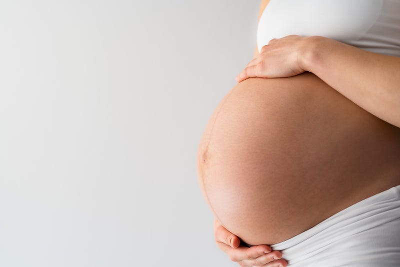 Description: Midsection of unrecognizable standing mother gently holding her very round pregnant baby belly. Side angle view. White background. Bright shot.