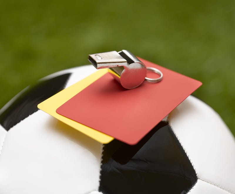 A yellow and red card resting atop a soccer ball with a referee's whistle.