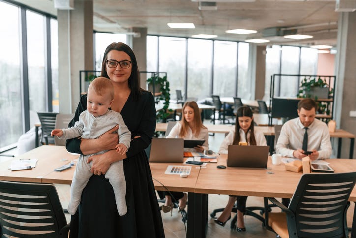 Happy mother is standing and holding her little son. Infant baby is in the office where group of people are working together.