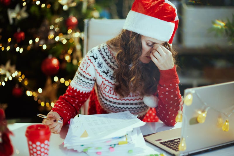 Christmas time. stressed stylish middle aged business woman in santa hat and red Christmas sweater with documents and laptop working in modern green office with Christmas tree.
