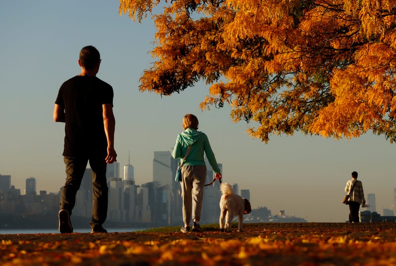 People walk along a walkway covered in fall foliage in Edgewater, New Jersey, as the sun rises over the New York skyline on Oct. 28, 2023, when highs hit 80 degrees in Central Park