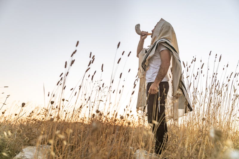 Jewish man in a traditional Tallit prayer shawl blowing the ram's horn Shofar, in the field against sunrise sky on Rosh Hashana and Yom Kippur.