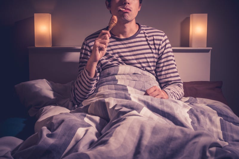 Young man is sitting in bed at night and eating a chicken drumstick