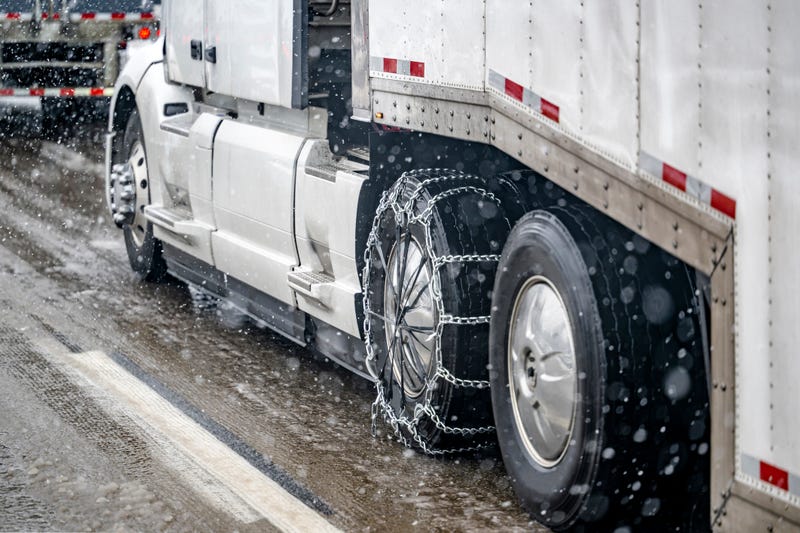 Required chains for better grip on slippery road surfaces on a white big rig semi truck drive axle wheel transporting cargo in dry van semi trailer running at winter weather during a snow storm