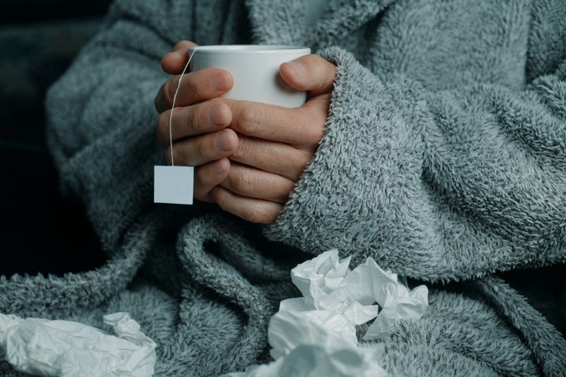 Ill man at home, wearing a fluffy gray house robe, surrounded by used tissues, warms up with a cup of hot herbal tea