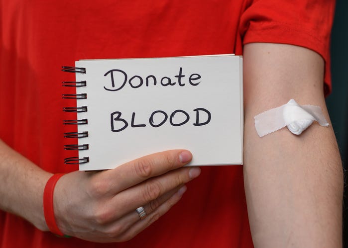  A voluntary blood donor wearing a red shirt, with a bandage and cotton ball over the bend of his elbow holds a sign that says "Donate blood"