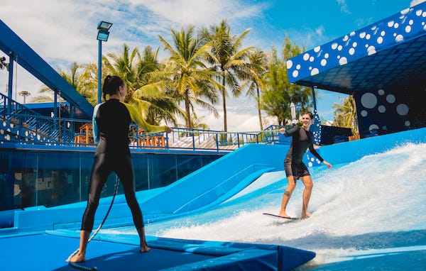 Young man surfing with trainer on a wave simulator at a water amusement park
