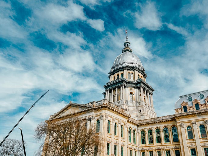 Rear views of the Illinois State Capitol Building in Springfield, Illinois, USA. Cloudy blue skies overhead. The US flag, State of Illinois Flag, and Black History Month Flag fly atop the dome. Construction crane in the view as construction work continues.