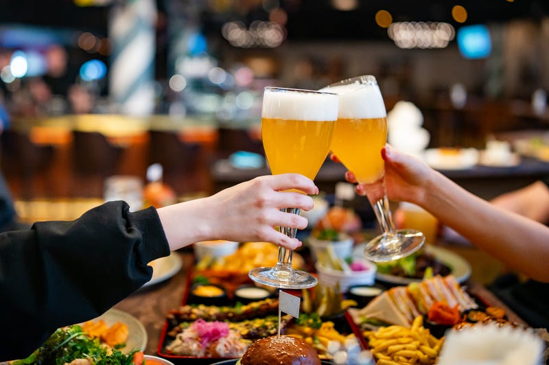 Two hands clinking beer glasses in a cozy indoor setting. Assorted food items are visible on a wooden table in the blurred background - stock photo