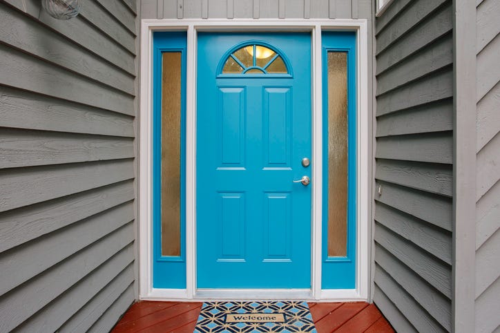 This image showcases a bright blue front door with two narrow side panels of frosted glass, a silver handle, and a welcome mat on a red wooden porch floor, surrounded by gray siding walls, creating a welcoming entrance to the home.