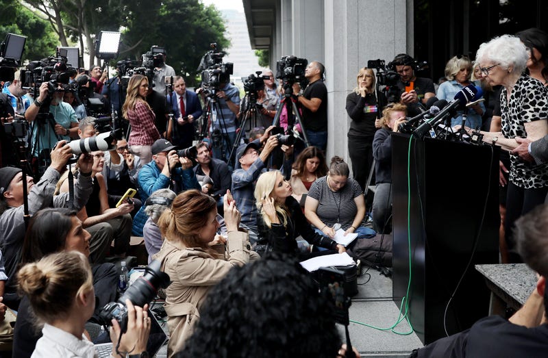elderly woman speaking in front of crowd of reporters
