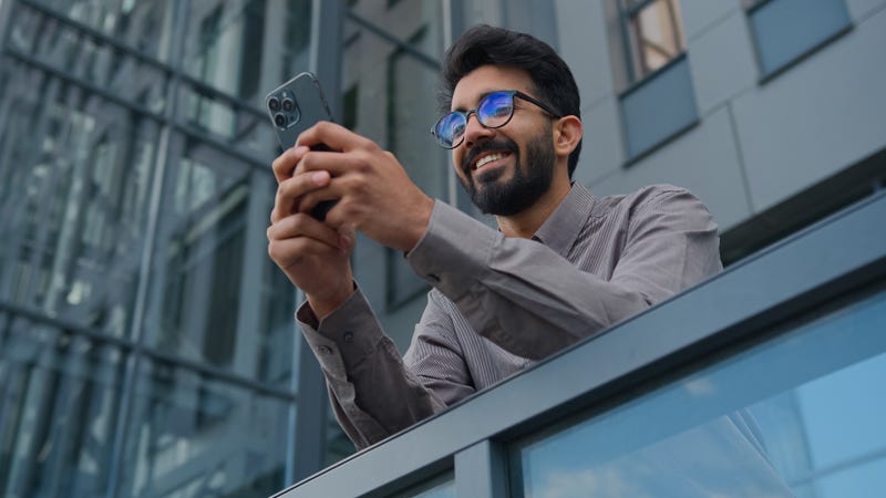 Smiling Arabian Indian man businessman entrepreneur manager use mobile phone application in city office terrace bottom view male business owner employer browsing social media texting cellphone message