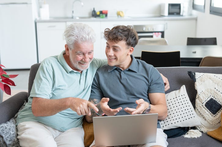 Happy multigenerational family sitting on sofa using laptop together - grandfather with young grandson enjoying carefree moments together learning use of computer