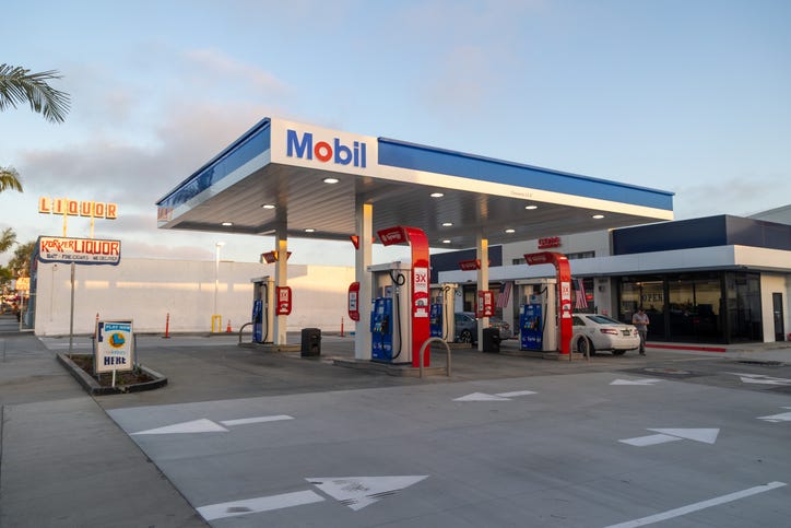A clean and modern Mobil gas station captured during golden hour in Newport Beach, California. The scene includes several fuel pumps under a brightly lit canopy, a convenience store, and classic signage from a nearby liquor store adding a nostalgic touch. Ideal for themes related to travel, fuel, transportation, road trips, and everyday life in Southern California.