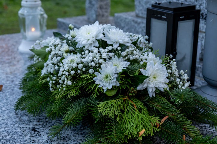 A cemetery bouquet made of fresh white chrysanthemum flowers rests on a gravestone next to a burning memorial candle. The soft glow of the flame and the purity of the white blossoms create a peaceful and respectful atmosphere, symbolizing love, remembrance, and eternal tribute to a departed loved one.
