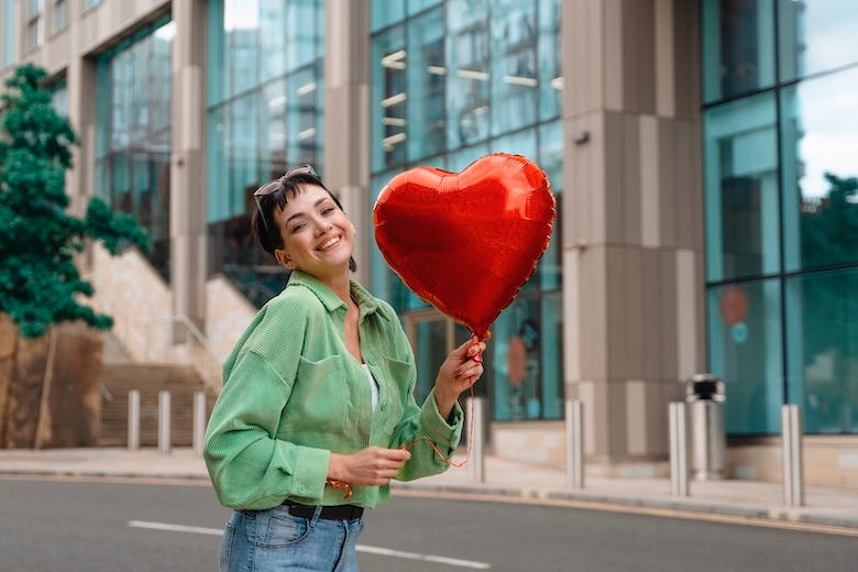 Woman smiles while holding red heart balloon on city street during daytime