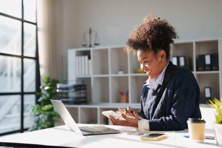 Smiling young adult woman concentrating on financial calculations using a calculator and laptop, working on business accounting and taxes in a professional environment