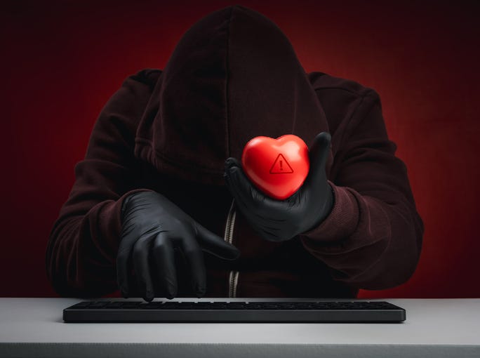Romance scam concept showing anonymous hacker in dark hoodie holding red heart with warning icon, symbolizing online love fraud, emotional manipulation, and cyber deception in dating scams.