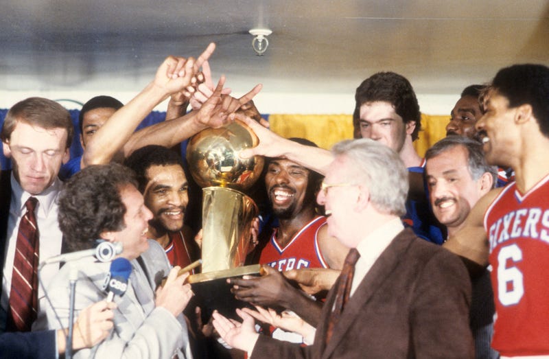 Philadelphia 76ers Maurice Cheeks (L) and Andrew Toney (R) victorious, holding Walter A. Brown trophy with teammates in locker room after winning game and series at The Forum. Game 4.