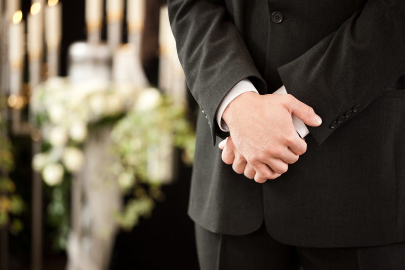 Funeral director stands at a service, an urn surrounded by flowers in the background