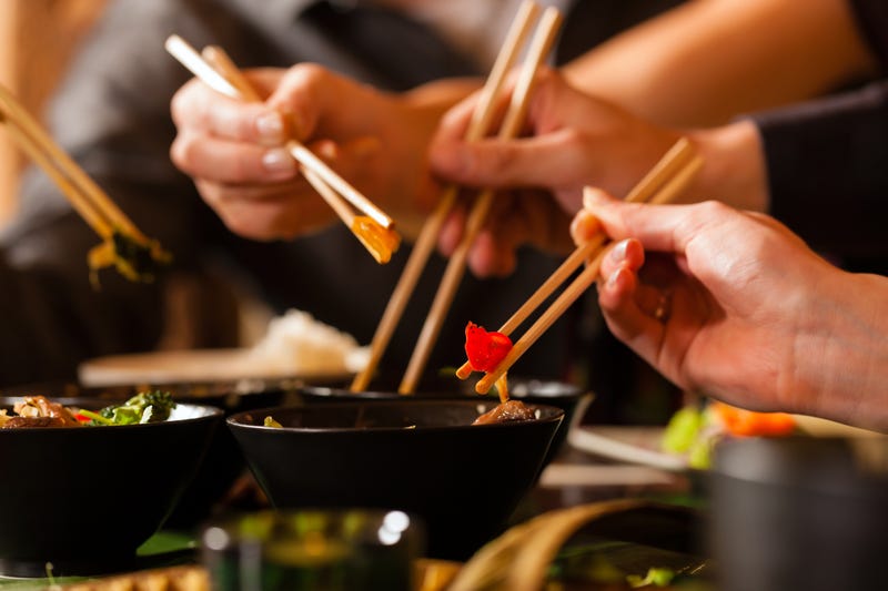 Four sets of chopsticks grabbing Asian food out of various black bowls sitting on a table in a Thai food restaurant.