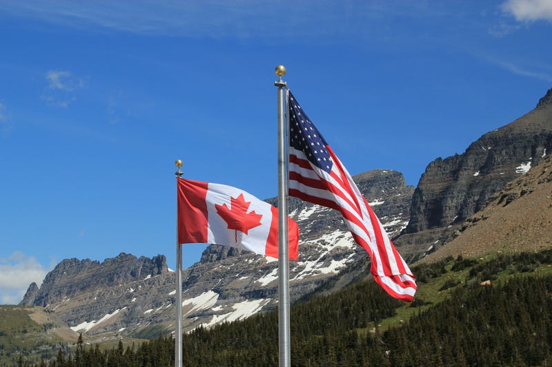 American and Canadian flags, showing the full stars and stripes and maple leaf respectively, on silver flagpoles blowing in the wind against a backdrop of snow covered mountains and forest trees