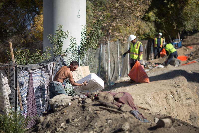 Rocky (L) cleans up the encampment of homeless friends who are faced with imminent eviction while they are away, as Cal Trans workers clean trash at a bridge construction site near the concreted channel of the Arroyo Seco Creek on November 20, 2015 in Los Angeles, California.